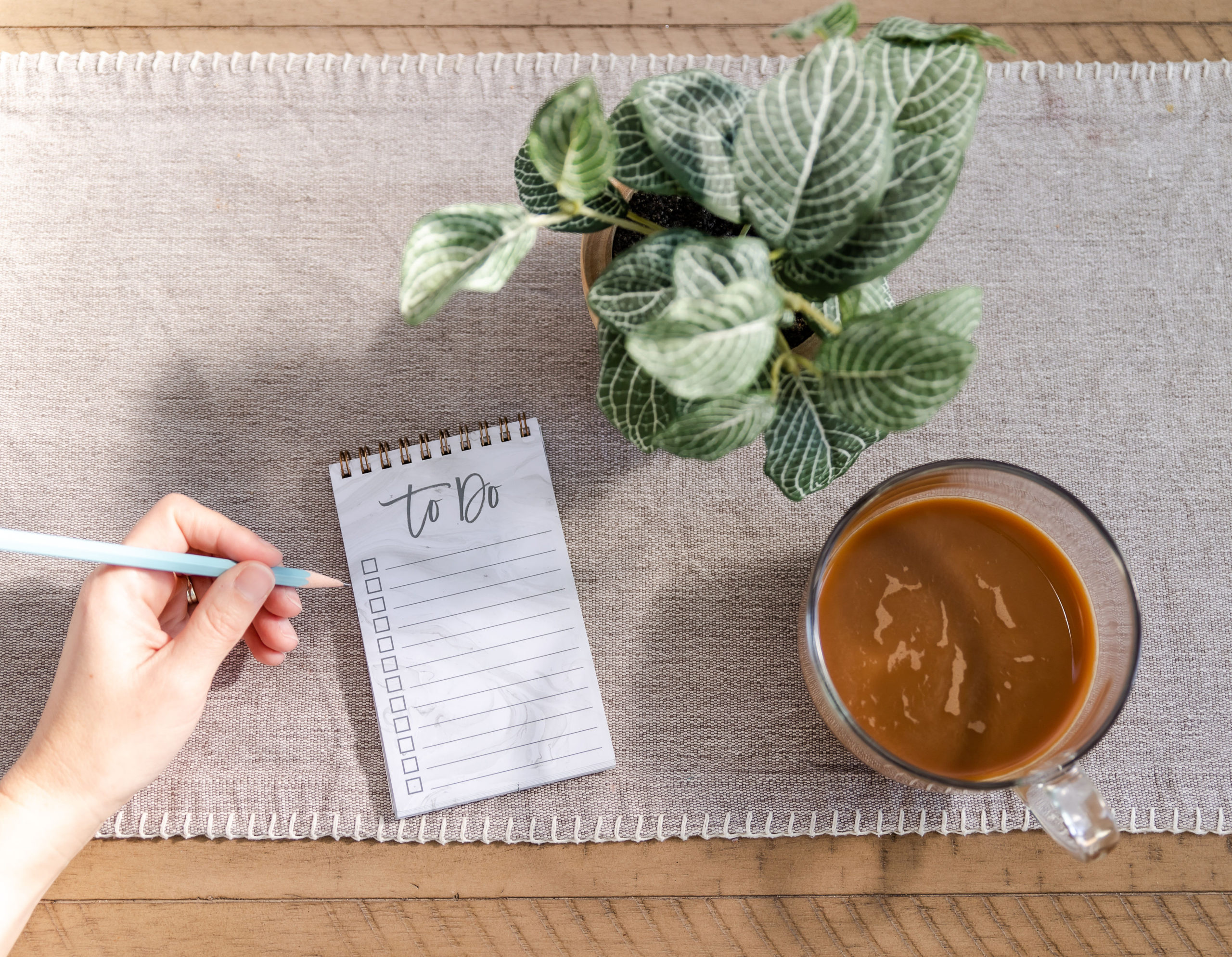 hand-holding-blue-pencil-writing-on-a-note-pad-to-do-with-glass-of-coffee-and-flower-pot-beside