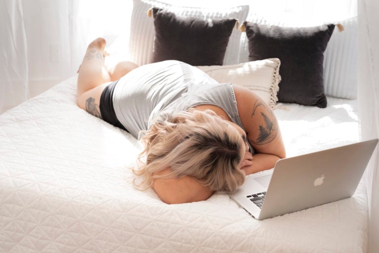 plus-size-blonde-hair-white-lady-lying-down-on-a-white-sheet-bed-with-open-laptop-on-the-bed