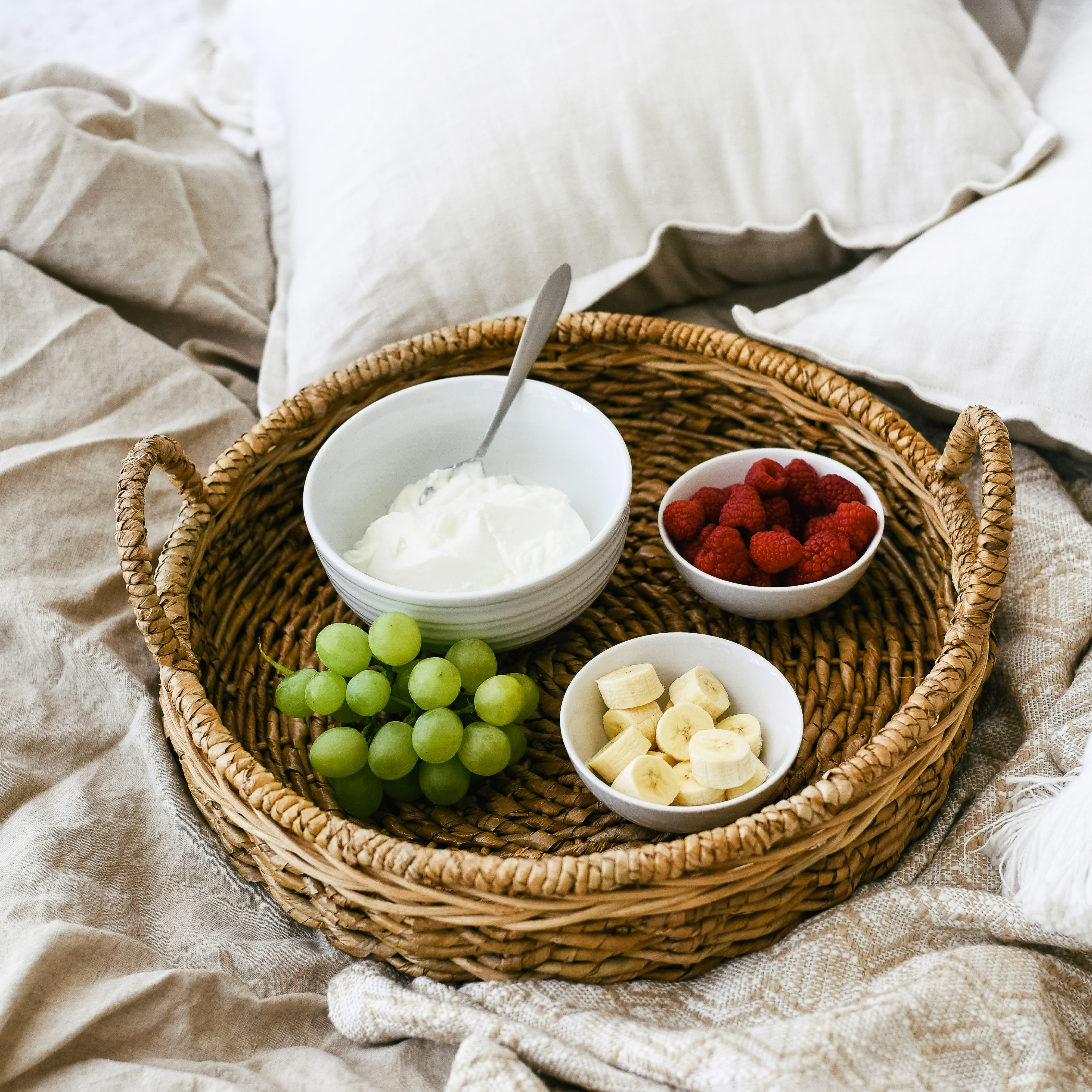 White-plate-grapes-sliced-banana-and-strawberry-in-a-round-basket-on-a-white-bed