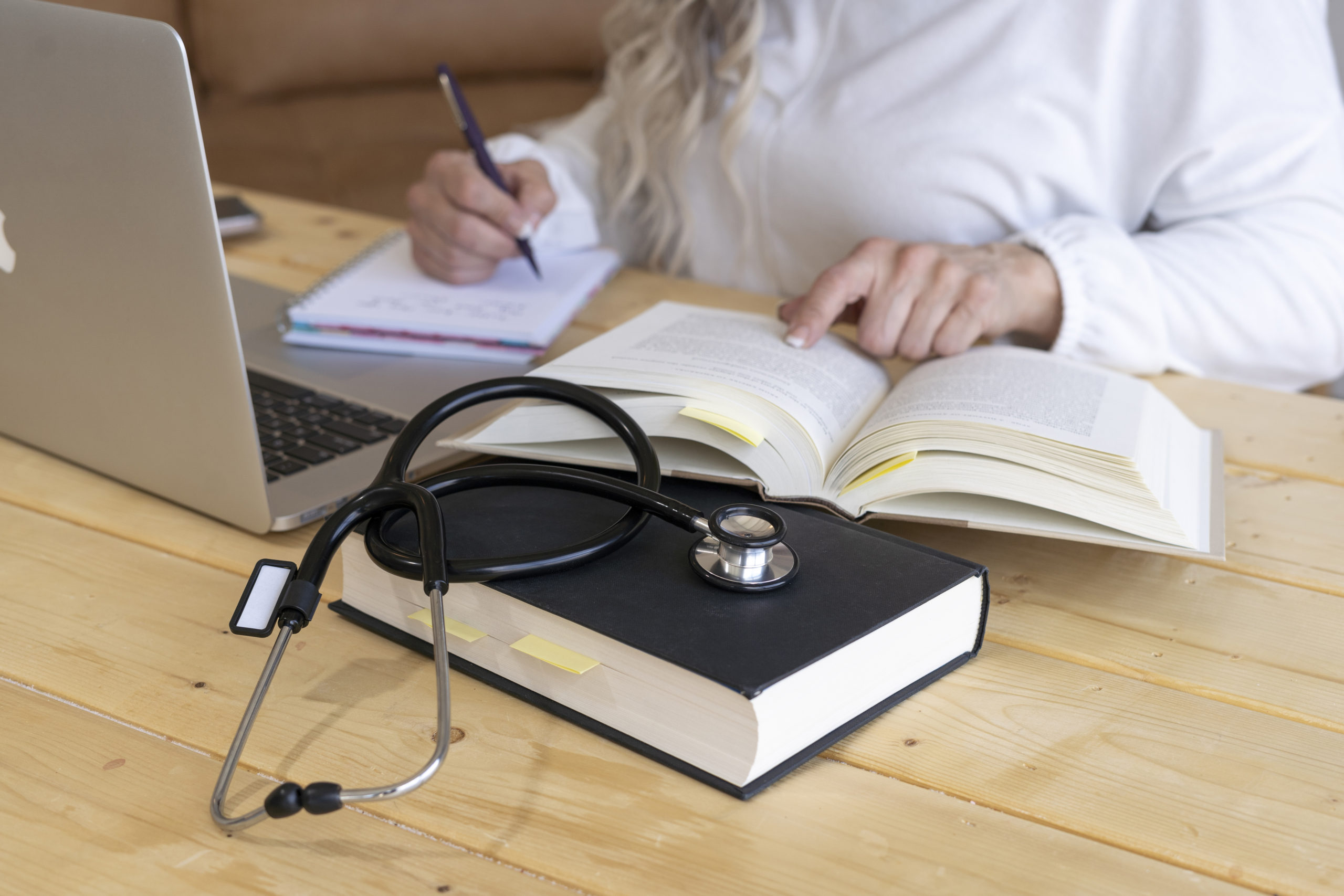 white-lady-reading-a-book-and-writing-on-a-brown-wooden-table-with-a-bible-and-stethoscope-beside