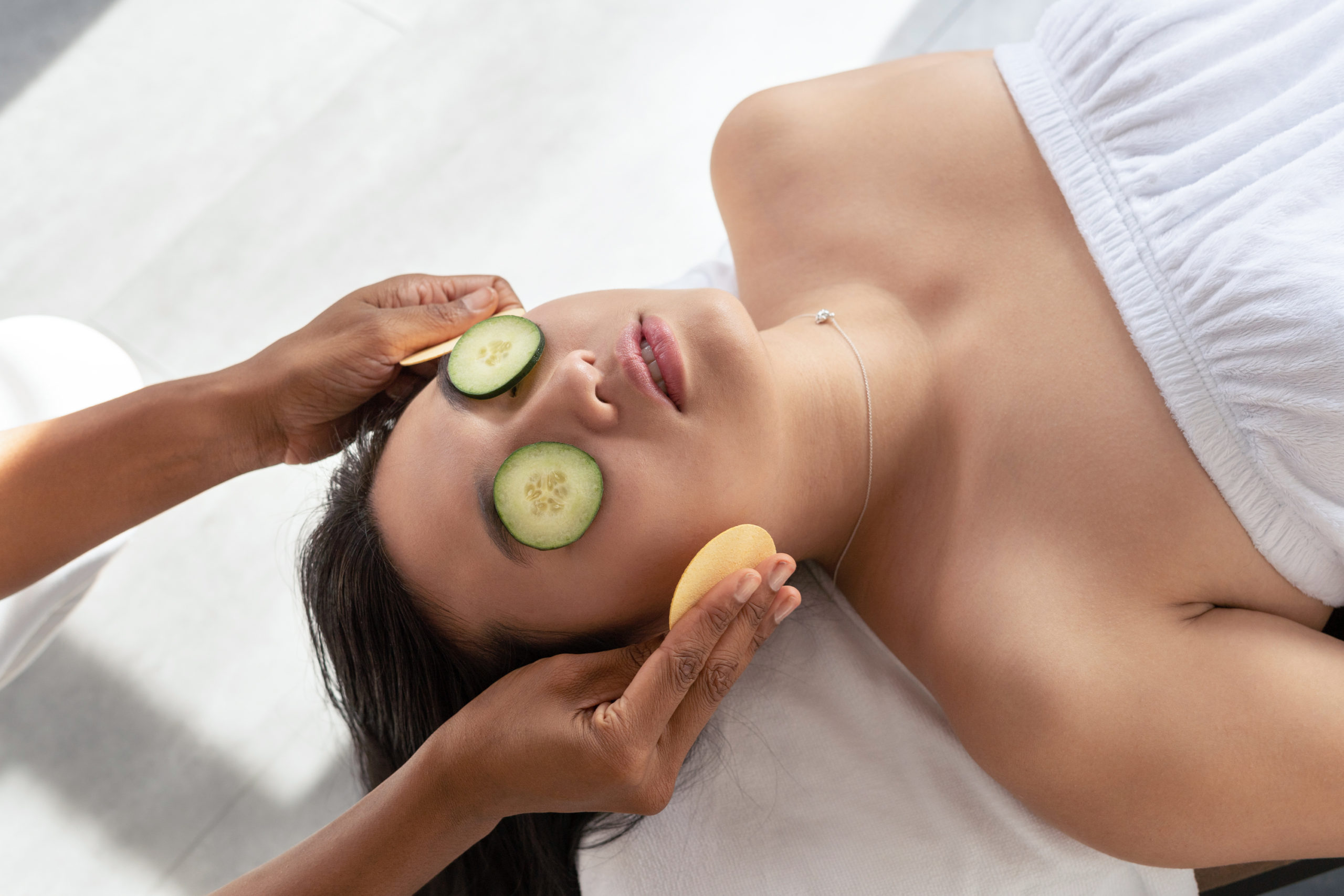 an-asian-lady-lying-on-her-back-wearing-a-white-towel-with-cucumber-slices-on-her-eyes-receiving-a-facial-massage