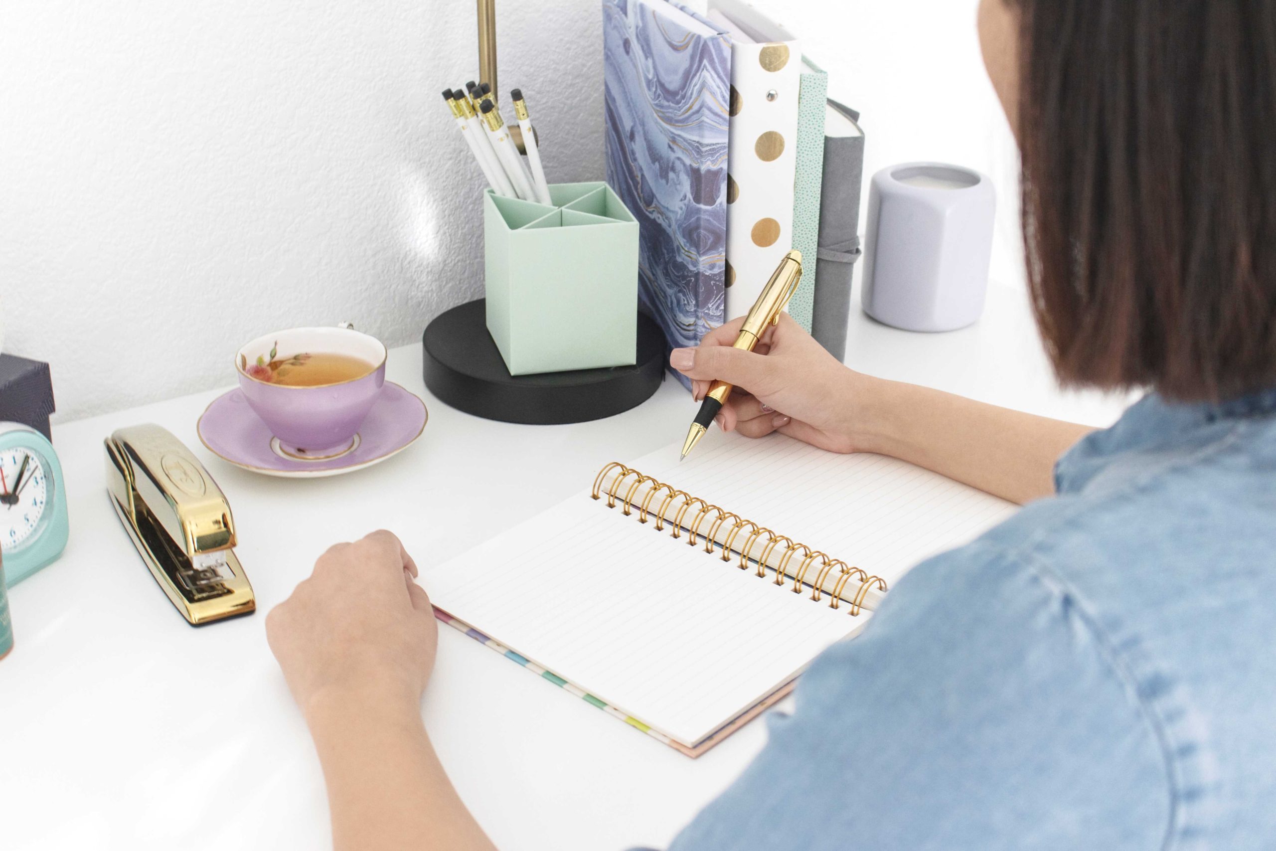 white lady wearing a sky blue denim shirt and writing with a gold pen on a note pad placed on a white table