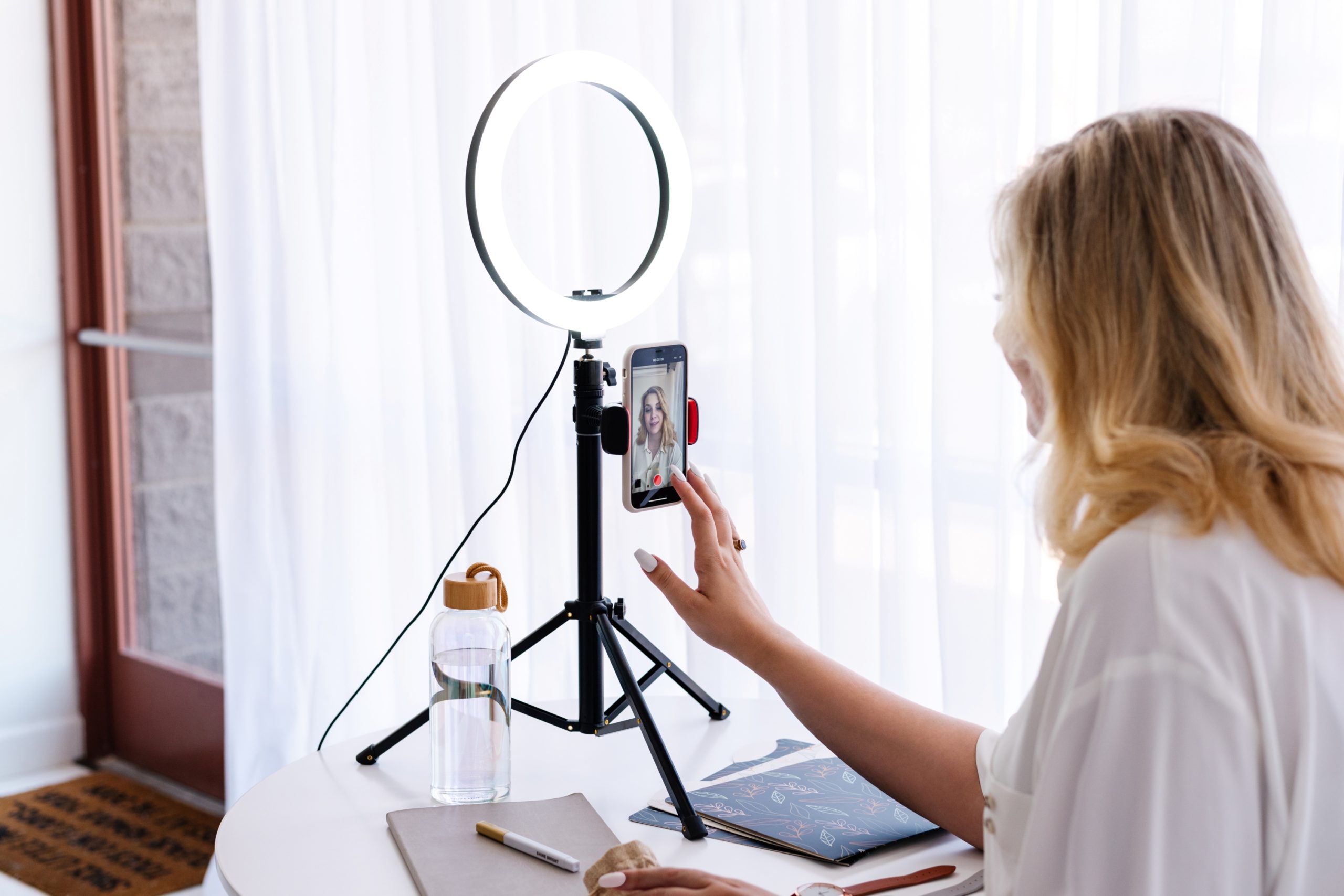 blonde lady wearing a white shirt and talking to a phone mounted on a ring light stand on a table