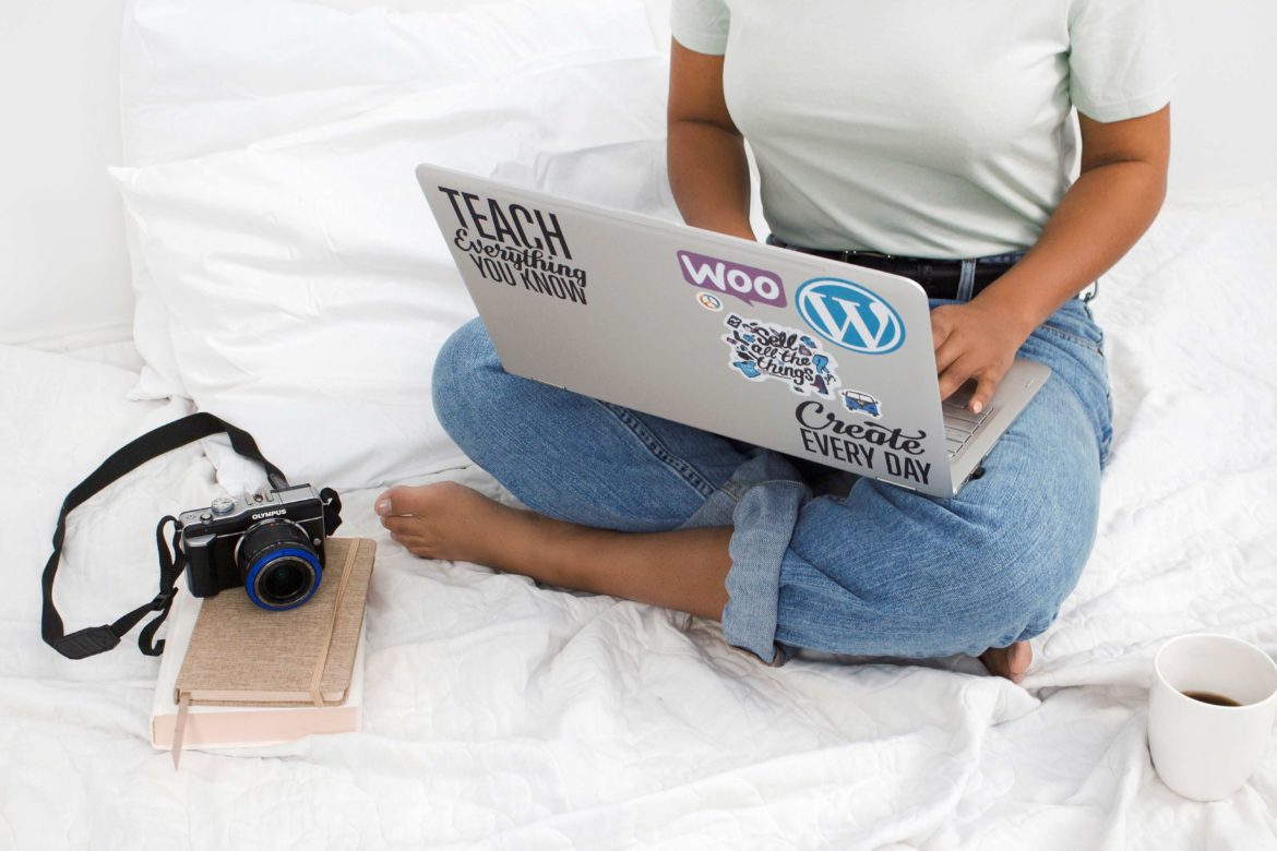 black lady wearing an ash tee shirt and a sky blue denim sitting on a white bed with a laptop on her laps with camera beside her