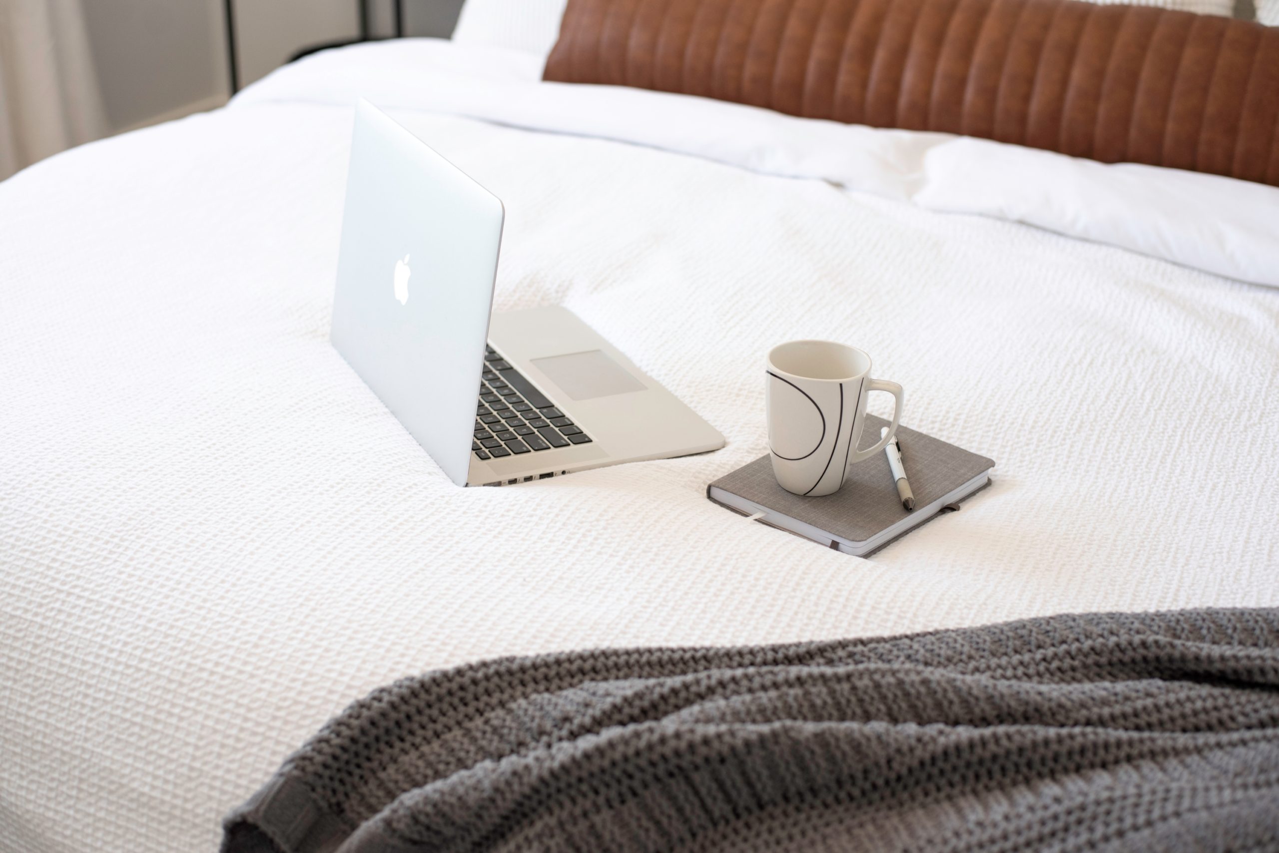 laptop journal and coffee mug on a white bed with gray comforter