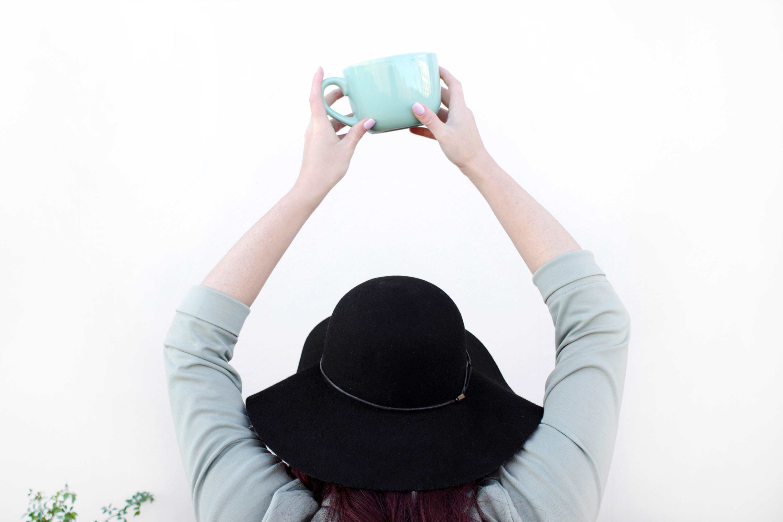 white lady wearing a dusty blue top and a black hat backing the camera and raising a sky blue cup up