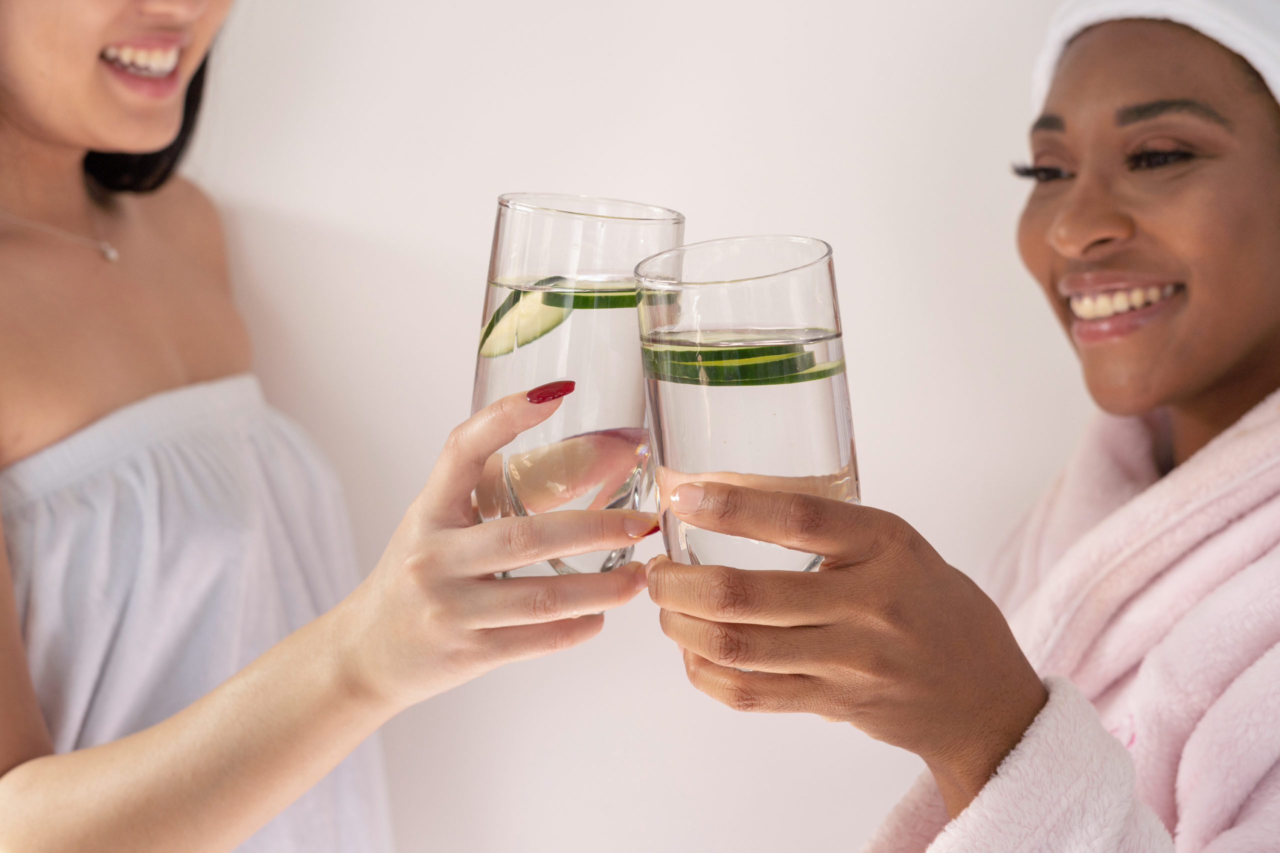 Two friends in spa mood laughing and making a toast with a glass of water with cucumber slices inside