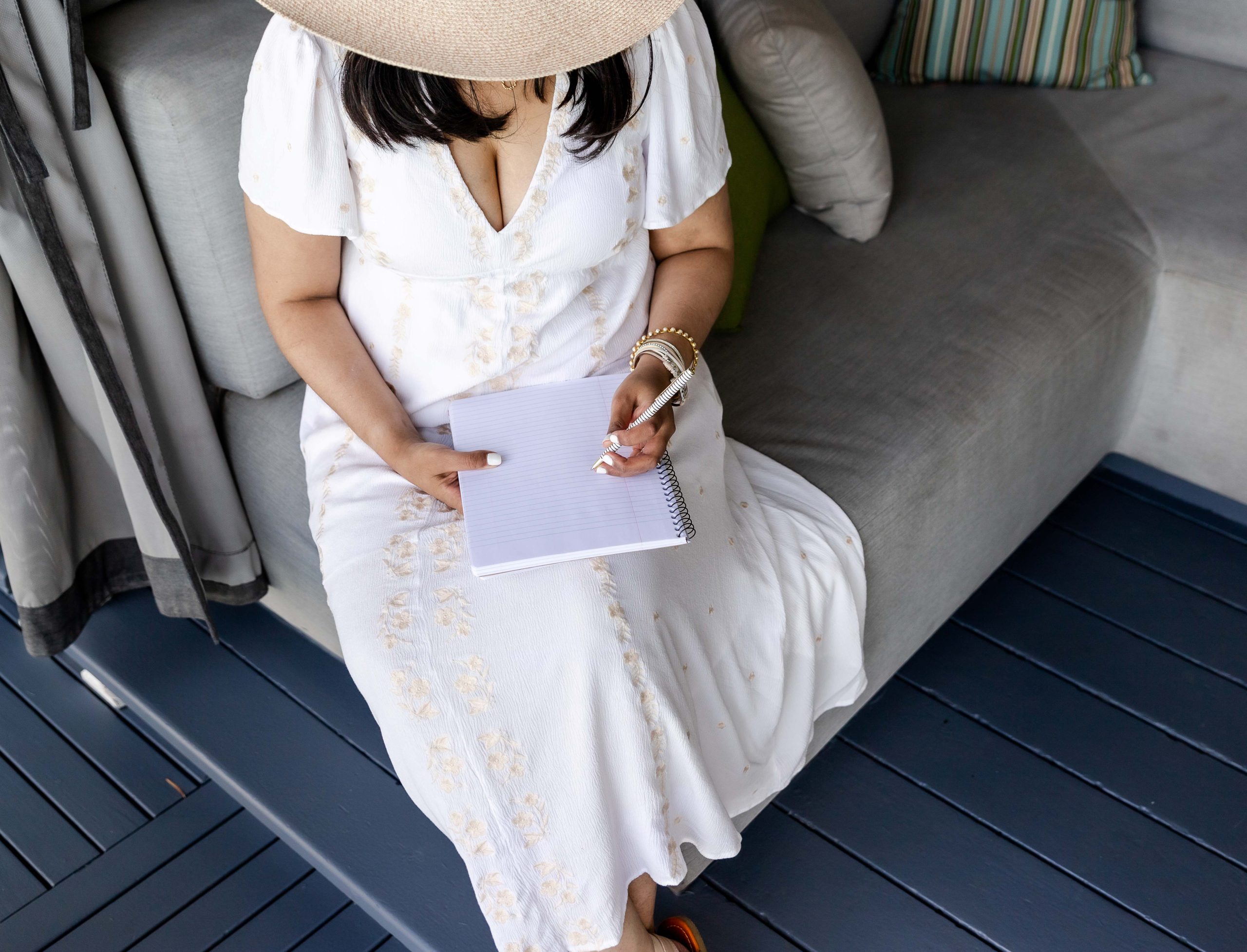 white woman wearing a white dress and a beach hat and writing on a white book while sitting on a couch
