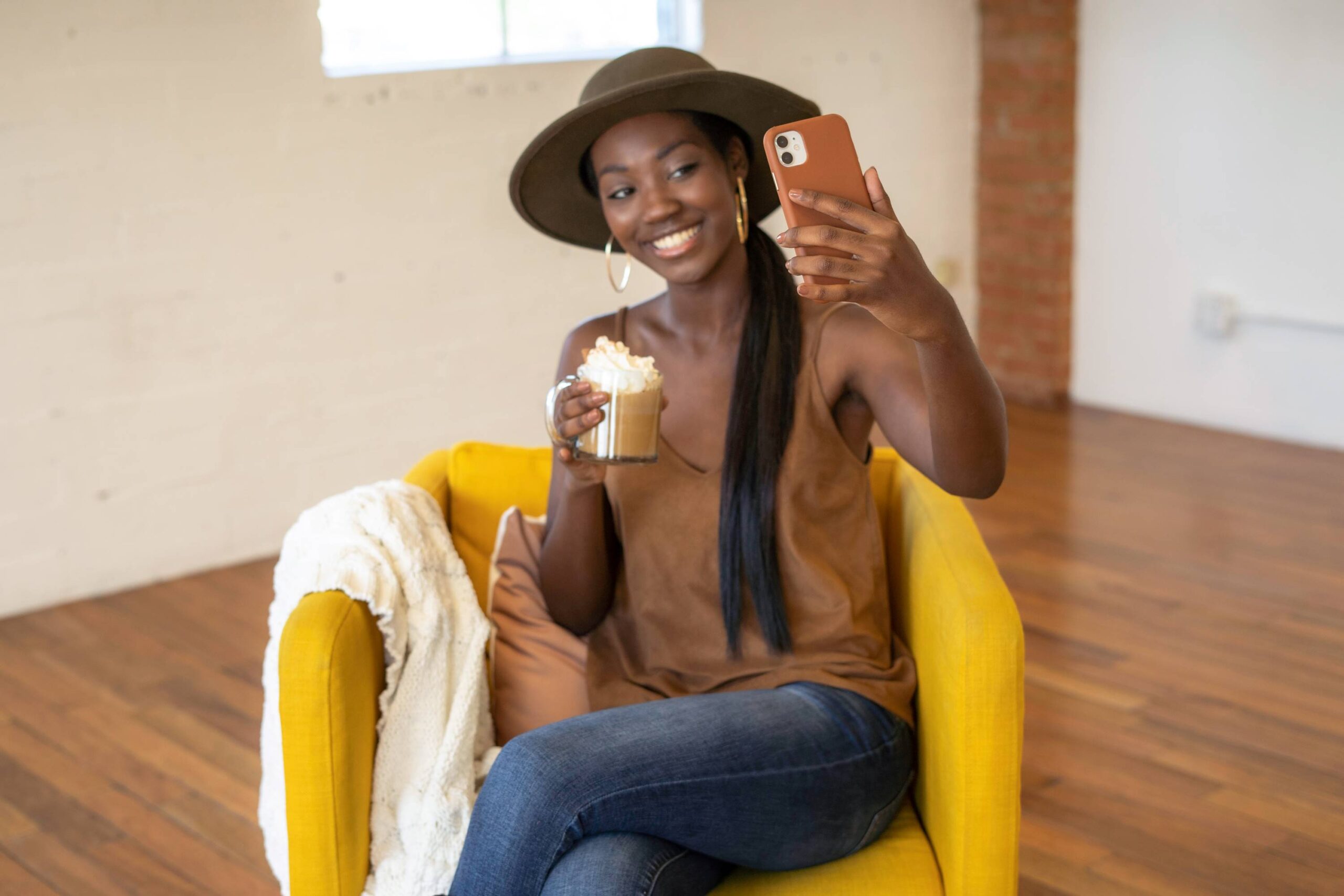black-lady-wearing-a-brown-fedora-hat-and-taking-a-selfie-while-sitting-on-a-yellow-chair